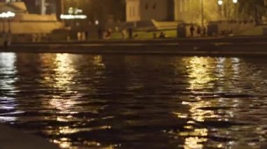 Suyun yüzeyini gece şehrinin ışıklarıyla kapatın. Stok görüntüleri. Yürüyen insanlarla dolu bir yaz gecesinde geniş bir nehir ve karanlık şehir caddesi dalgalandı..