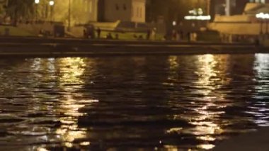 Suyun yüzeyini gece şehrinin ışıklarıyla kapatın. Stok görüntüleri. Yürüyen insanlarla dolu bir yaz gecesinde geniş bir nehir ve karanlık şehir caddesi dalgalandı..