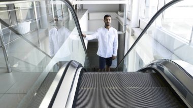 Beyaz tişörtlü bir adam iş merkezinin içinde yürüyen merdivenden çıkıyor. HDR. Mağazadaki yürüyen merdivendeki genç adam..