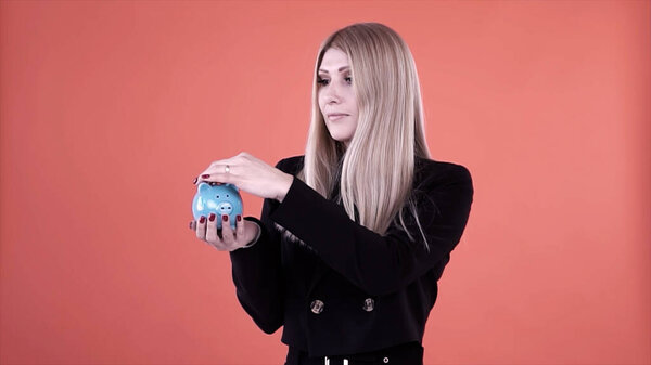 Blond female putting coin into a blue piggy moneybox isolated on pink background. Clip. Serious female in black jacket making savings, concept of investing money and financial economy.