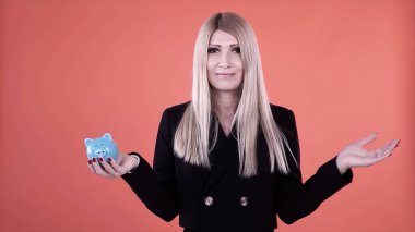 Sarışın kadın, pembe arka planda izole edilmiş mavi bir para kutusuna bozuk para koyuyor. Şarjör. Siyah ceketli ciddi bir kadın tasarruf yapıyor, para yatırımı ve finansal ekonomi kavramı.