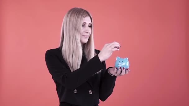 Blond female putting coin into a blue piggy moneybox isolated on pink background. Clip. Serious female in black jacket making savings, concept of investing money and financial economy. — Stock Video