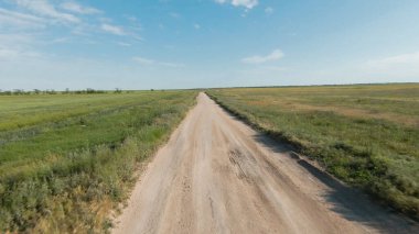 Tarımsal alanların yakınındaki kırsal yolun en üst görüntüsü. Vuruldu. Ufkun arka planında mavi gökyüzü ile birlikte insansız hava aracı yol üzerinde uçuyor. Güneşli bir günde yol ve yeşil tarlalarla güzel bir manzara.