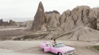 Türkiye, Kapadokya 'daki fotoekonomik durgunluk döneminde genç ve güzel bir beyaz gelin ve damat. Başla. Kaya oluşumlarının arka planında pembe renkli bir Cabriolet arabada yeni evli bir çiftin hava görüntüsü..