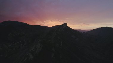 Mor günbatımı gökyüzünün arkaplanındaki dağın üst manzarası. Başla. Arka plandaki karanlık dağ siluetleri günbatımı gökyüzünde. Karanlık dağların tepelerinde kasvetli gökyüzü