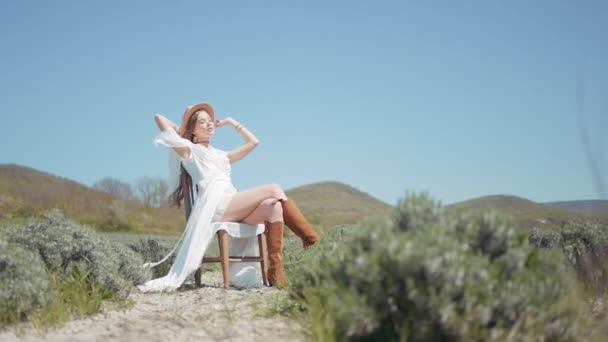 En vacker ung kvinna njuter av solig sommardag på ett lavendelfält. Börja. Kvinna med långt hår i en vit klänning, cowboy stövlar och en hatt sitter i en stol bland blommande blommor på blå klar himmel — Stockvideo