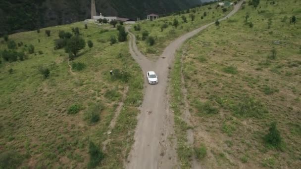 Luchtfoto van een witte personenauto op een landelijk smal weggetje in de bergen. Actie. Een voertuig dat langs zeldzame bomen en groene hellingen rijdt. — Stockvideo