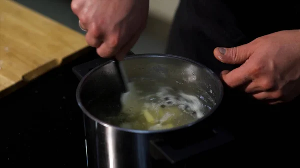 Närbild av tillagning av soppa, buljong med potatis och svartpeppar korn. Konst. Chef händer under omrörning soppa med ett kök spatel. — Stockfoto