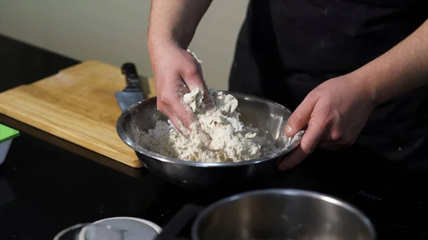 Mannen knådar deg med händerna i en metallskål i köket. Konst. En kock förbereder degen, blanda ingredienser med händerna. — Stockfoto