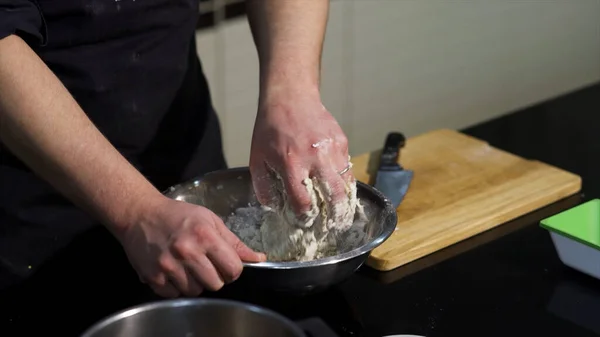 Mannen knådar deg med händerna i en metallskål i köket. Konst. En kock förbereder degen, blanda ingredienser med händerna. — Stockfoto