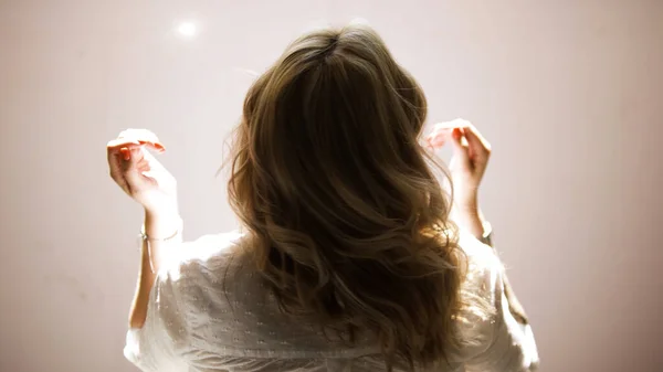 Egy fiatal nő rázza a szőke göndör haját elszigetelve egy bézs fal hátterében. Művészet. Hátsó nézet egy fiatal szőke nő fehér ingben pózol és megérinti a haját. — Stock Fotó