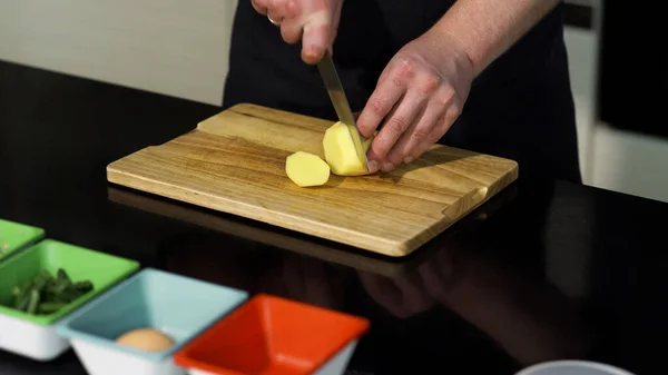 Un uomo che taglia una patata pelata su una tavola di legno. Art. Primo piano dello chef maschile che prepara un piatto e utilizza un coltello d'acciaio per tagliare le verdure. — Foto Stock