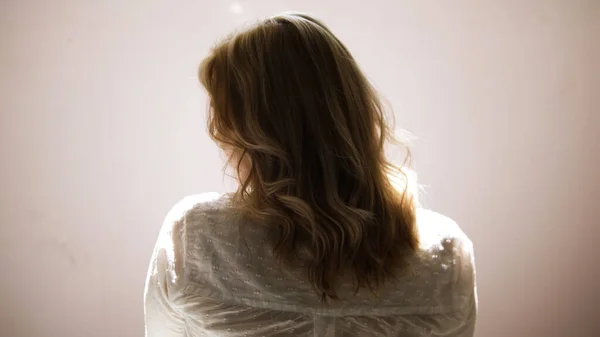 Una joven sacudiendo su rubio pelo rizado aislado sobre un fondo de pared beige. Art. Vista trasera de una joven rubia con una camisa blanca posando y tocándose el pelo. — Foto de Stock