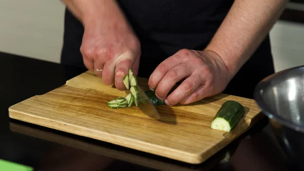 Un uomo che taglia un cetriolo pelato su una tavola di legno. Art. Primo piano dello chef maschile che prepara un piatto e utilizza un coltello d'acciaio per tagliare le verdure. — Foto Stock