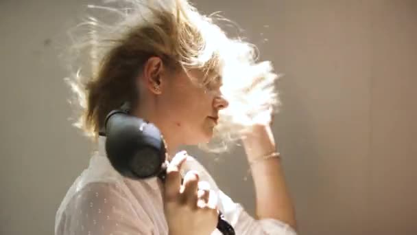 Mladá žena si suší vlasy fénem izolovaným na béžovém pozadí. Umění. Mladá blondýnka v bílé košili styling ji vlasy. — Stock video
