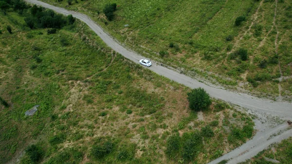 Widok z lotu ptaka na biały samochód pasażerski jadący wąską drogą w górach. Akcja. Pojazd poruszający się wzdłuż rzadkich drzew i zielonych zboczy wzgórz. — Zdjęcie stockowe