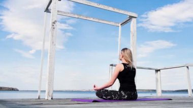 Sarışın genç bir kadın göl kenarında nilüfer pozisyonunda oturuyor ve nefes alma egzersizleri yapıyor. Kavram. Spor takım elbiseli bir kız mavi bulutlu arka planda bir iskelede meditasyonda oturuyor..