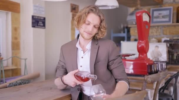 Un bel homme aux cheveux blonds bouclés en costume beige versant du vin de rose dans un verre transparent. L'action. Sommelier masculin versant du vin de fruits dans un bar. — Video