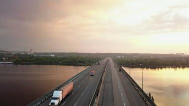 Alacakaranlıkta geniş bir nehri kapsayan uzun bir otoyol köprüsünde giden araba ve kamyonların hava görüntüsü. Kırp