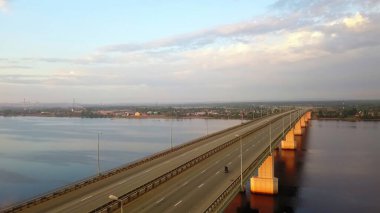 Beautiful aerial view showing a motorcyclist riding across a long bridge spanning a river at sunrise. Clip