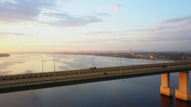 Aerial view of a concrete bridge over a calm river with city traffic moving at sunset. Clip