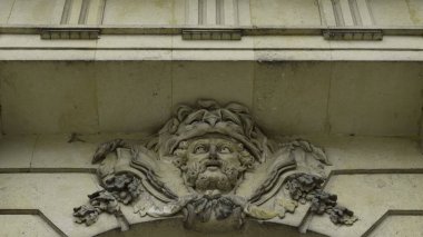 Yapraklı bir taç ve örtülü kumaşla taş maskarası, bir binanın ön cephesinde mimari bir detay. Eylem