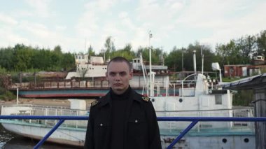 Kaptan üniformalı genç adam arkasında paslı gemilerle kameraya ciddi ciddi bakıyor. Kırp