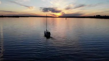 Renkli bir günbatımında sakin sularda süzülen yelkenli sakin yüzeyi yansıtıyor.