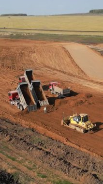 Çöp kamyonları inşaat alanına toprak boşaltıyor ve bir buldozer zemini yerle bir ediyor.