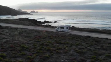 Güzel bir gün batımında okyanusun yanındaki sahil yolunda giden bir karavanın hava görüntüsü. Medya