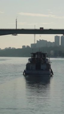 Eski römorkör beton bir köprünün altındaki geniş bir nehirde yol alırken şehir trafiği ve arkadaki ufuk çizgisi. Kırp