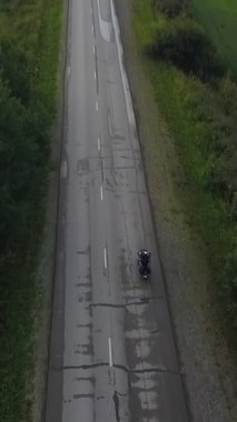 Şehre doğru yeşil tarlalardan geçen uzun kırsal bir yolda giden bir motosikletlinin hava görüntüsü. Kırp
