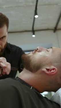 Odaklanmış berber müşterinin sakalını dikkatlice tıraş ediyor ve boyun hizasını usturayla şekillendiriyor. Kırp