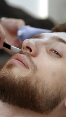 Kozmetik uzmanı genç sakallı bir adamın yüzüne nemlendirici bir maske takıyor. Kırp