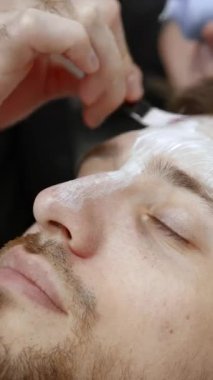 Kozmetik uzmanı, genç bir adamın alnına fırçayla kozmetik nemlendirici krem maske uyguluyor. Kırp