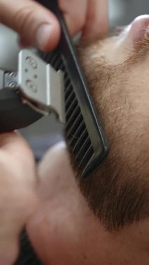 Genç bir adamın sakalını titizlikle şekillendirmek için saç kesme makinesi ve tarak kullanan bir berbere yakından bakın. Kırp