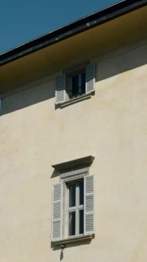 Zarif taş pencere çerçeveleri ve kepenkleri olan krem renkli bina cephesi, açık mavi gökyüzüne ve yeşil tepelere karşı kurulmuş.