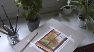 Pencere kenarındaki suluboya tablo, kavanozdaki fırçalar, maskeli bant kağıdı, saksı bitkileri çerçevesi. Medya