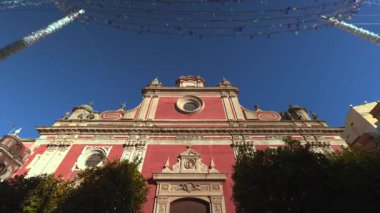 İp ışıklarının altındaki pembe kilise cephesi, süslü giriş ve ağaçlarla çerçevelenmiş Rosette penceresi, şenlikli tepe örtüsü. Eylem