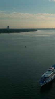 Beyaz ve mavi yolcu feribotu gün doğumunda bir nehirde yüzüyor, şehir manzarası ve güneş suları yansıtıyor. Kırp