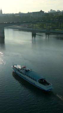 Beyaz yolcu feribotu gün doğumunda bir nehirde yol alırken, arka planda bir köprü ve şehir silüeti var. Kırp