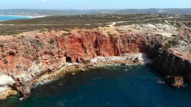 Hava kırmızı uçurum kıyısı kıvrımlı toprak yolu, turkuaz su, engebeli çalılık, panoramik ufuk, güneş ışığı