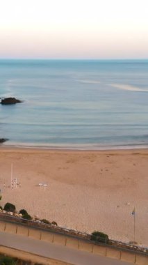 Şafak vakti kumlu kumsalda yumuşak dalgalar ve geniş körfez, yumuşak pastel gökyüzü, sessiz gezinti alanı ve park edilmiş arabalar.