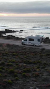 Güzel bir gün batımında okyanusun yanındaki sahil yolunda giden bir karavanın hava görüntüsü. Medya