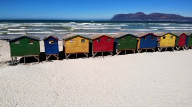 Colorful Beach Huts On White Sand Lined In Vibrant Wooden Cabins On Stilts, Rolling Ocean Waves Under Clear. Media