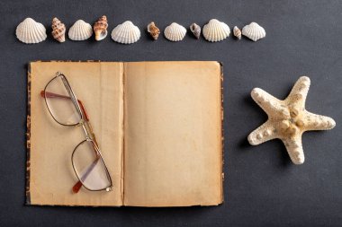 Eski sarı defter, gözlük ve deniz kabukları. Tatil anılarını saklayabileceğin bir yer. Koyu arkaplan.