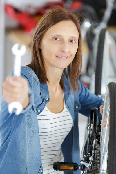 日当たりの良いガレージで自転車のワイヤーをチェックする美しい陽気な女性 — ストック写真