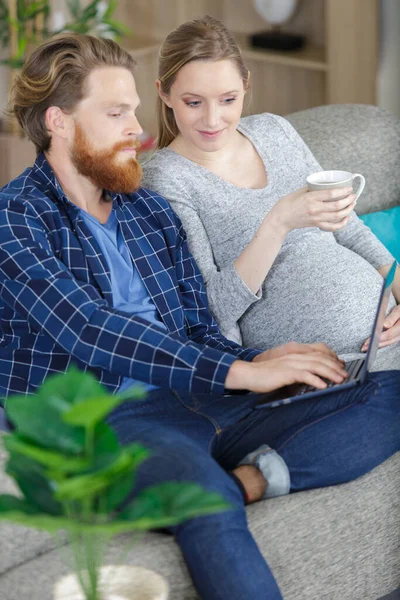 这对怀孕夫妇正在使用笔记本电脑在沙发上放松 — 图库照片