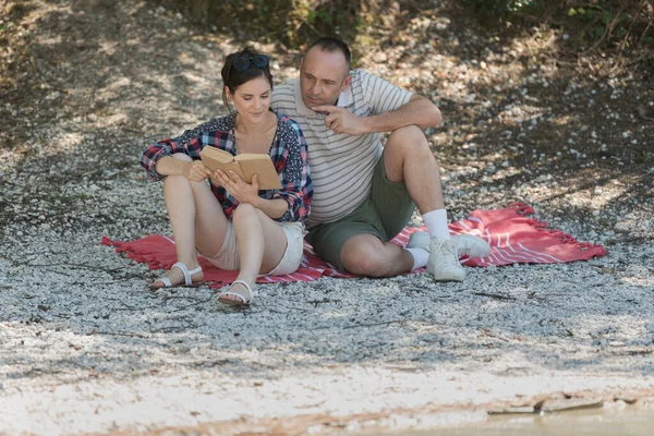 Coppia Guardando Libro Seduto Sulla Coperta Sulla Riva Del Fiume — Foto Stock