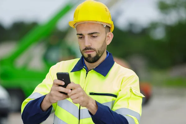 Uśmiechnięty Młody Człowiek Budowniczy Twardym Kapeluszu Pomocą Telefonu Komórkowego — Zdjęcie stockowe